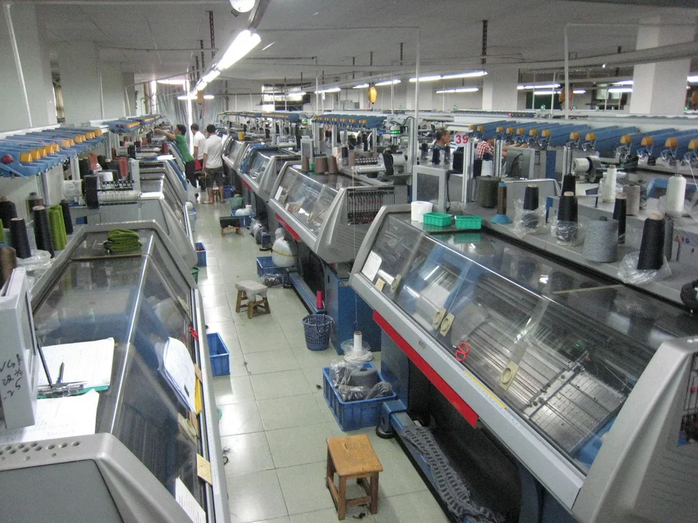 Rows of flat bed knitting machines with yarn cones and transparent covers